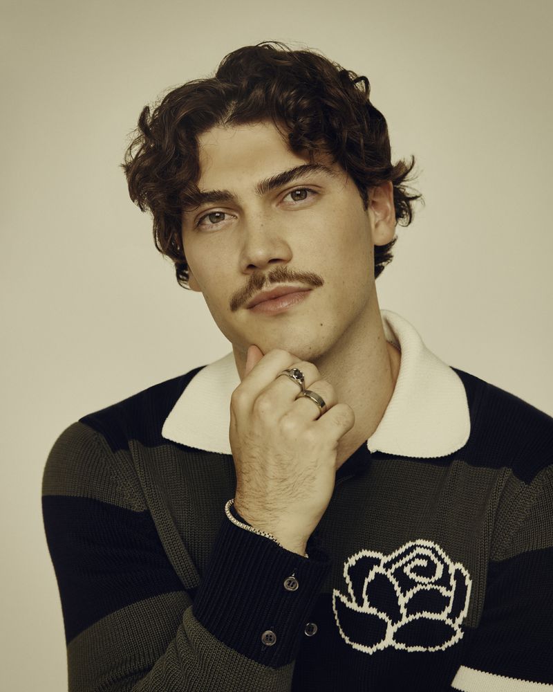 Portrait of a young adult man with curly hair and a mustache, wearing a striped sweater with a rose design, posing indoors.