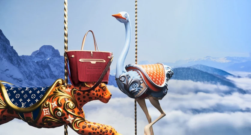 Louis Vuitton holiday campaign featuring sculpted animal carousel figures and a red handbag against a snowy mountain backdrop.