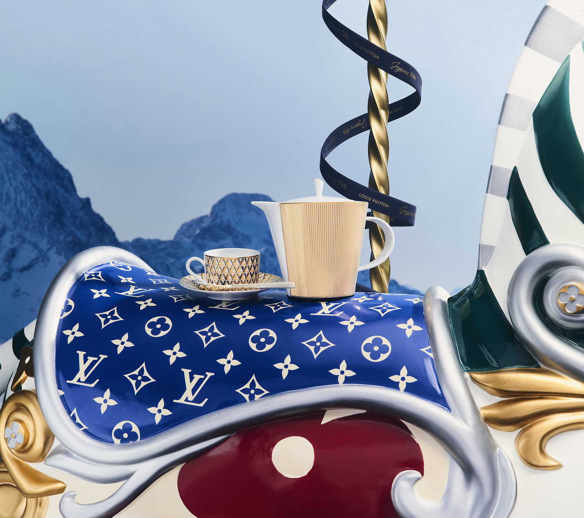 Porcelain teapot and teacup resting on a blue monogram saddle atop a decorative carousel against mountain backdrop