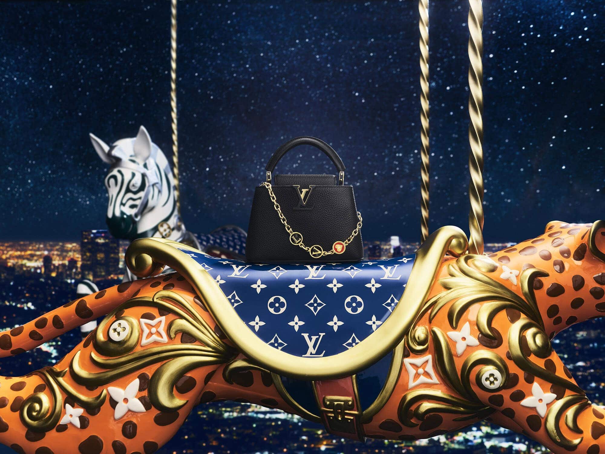 Black leather handbag with gold chain resting on a carousel animal against a starry night cityscape.