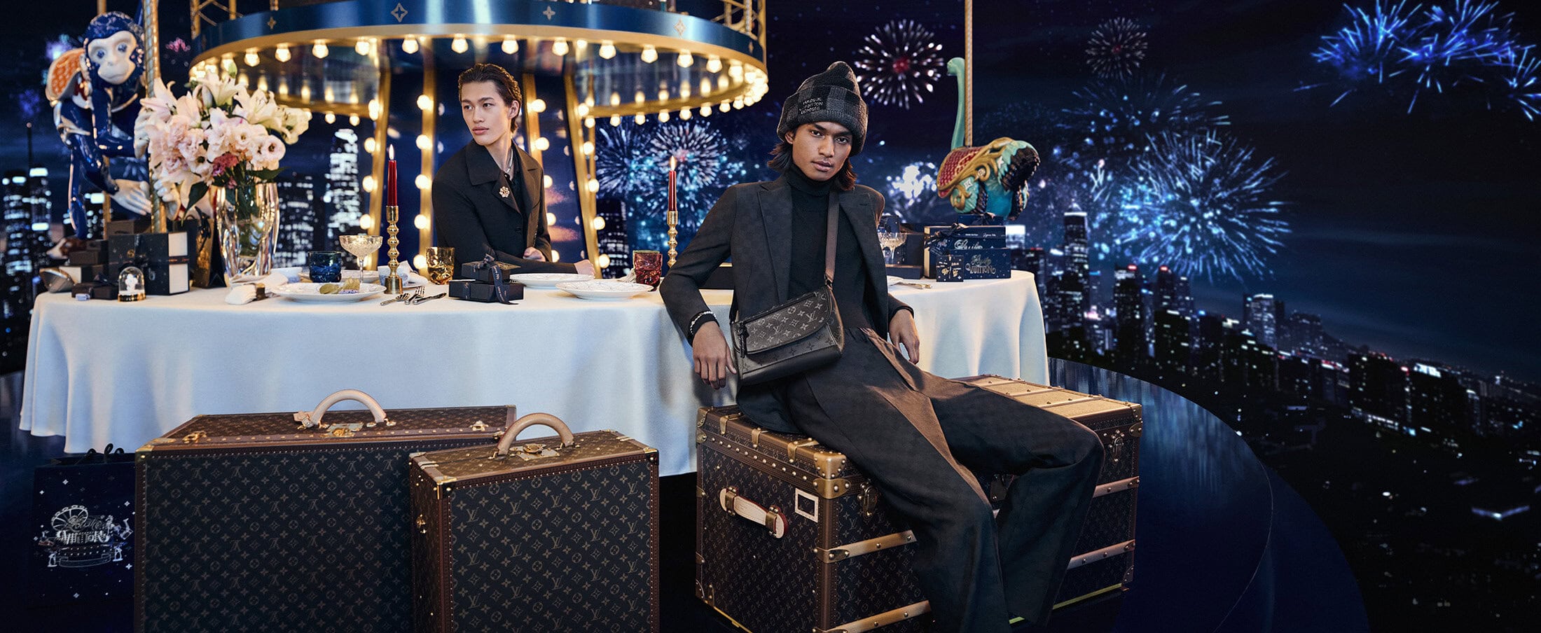 Model seated on luxury monogram trunks wearing a dark suit and beanie in front of a festive banquet table under a carousel and fireworks
