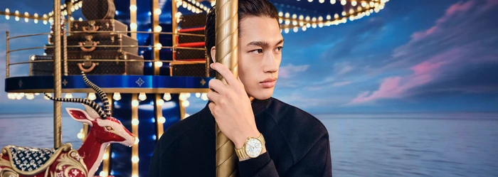 Young man holding a carousel pole wearing a dark jacket with a gold watch against a carousel and sunset sky