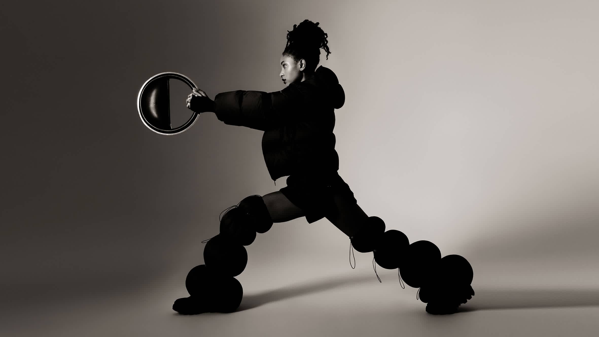 Willow Smith in a black puffer jacket and sculptural trousers holding a round ring prop against a grey studio backdrop.