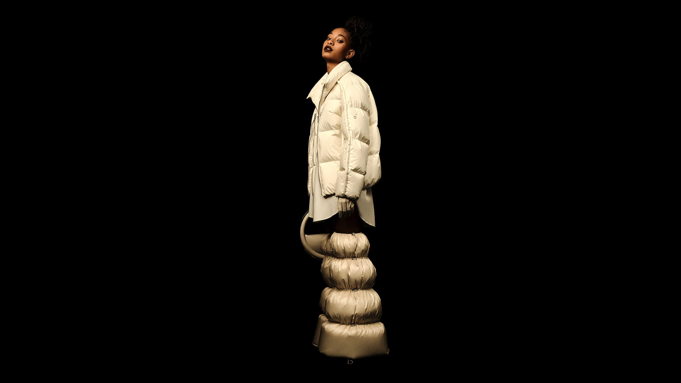 Model Willow Smith wearing a white puffer jacket and puffy boots against a black background for Moncler