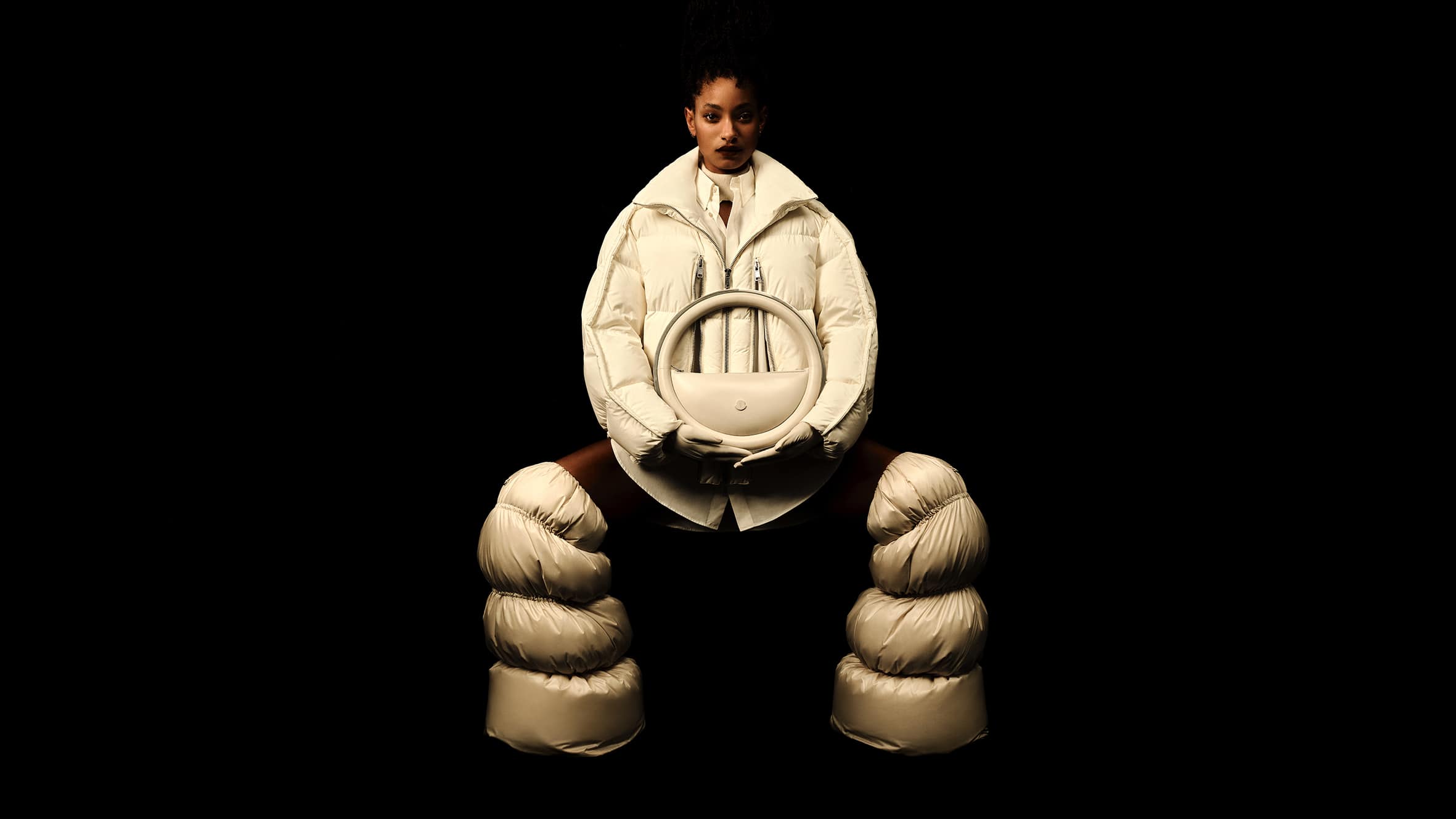Willow Smith wearing Moncler white puffer outerwear and quilted boots against a black background