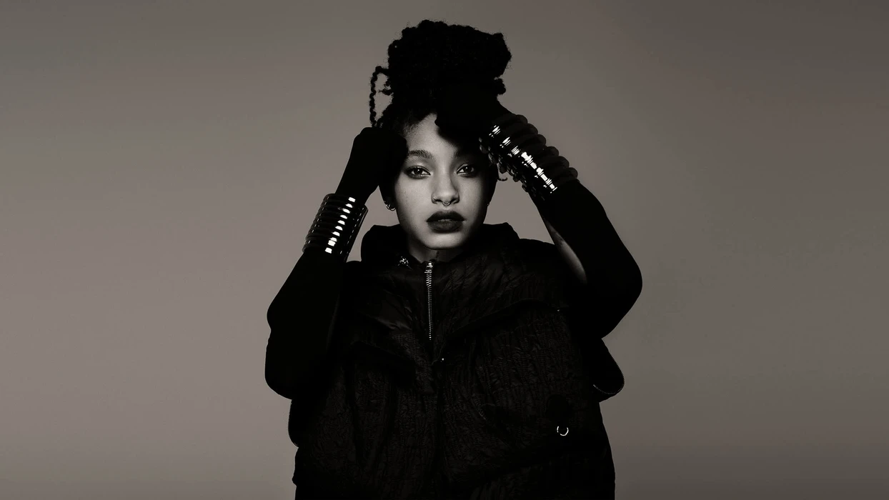Monochrome fashion portrait of Willow Smith wearing a dark puffer vest and metallic wrist cuffs against a grey studio backdrop.