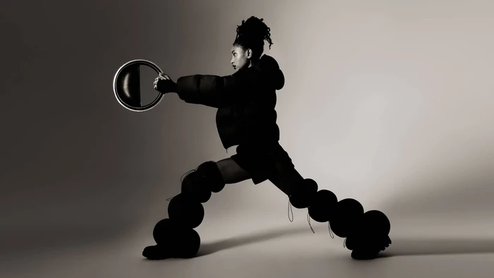 Willow Smith in a black puffer jacket and sculptural trousers holding a round ring prop against a grey studio backdrop.