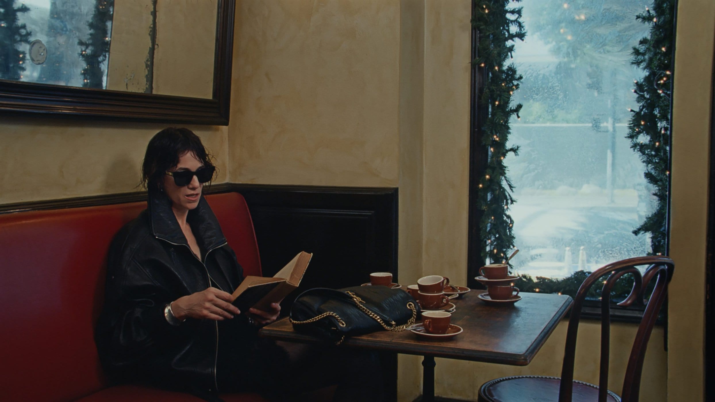Woman wearing dark sunglasses and a black leather jacket reading a book at a café table with teacups