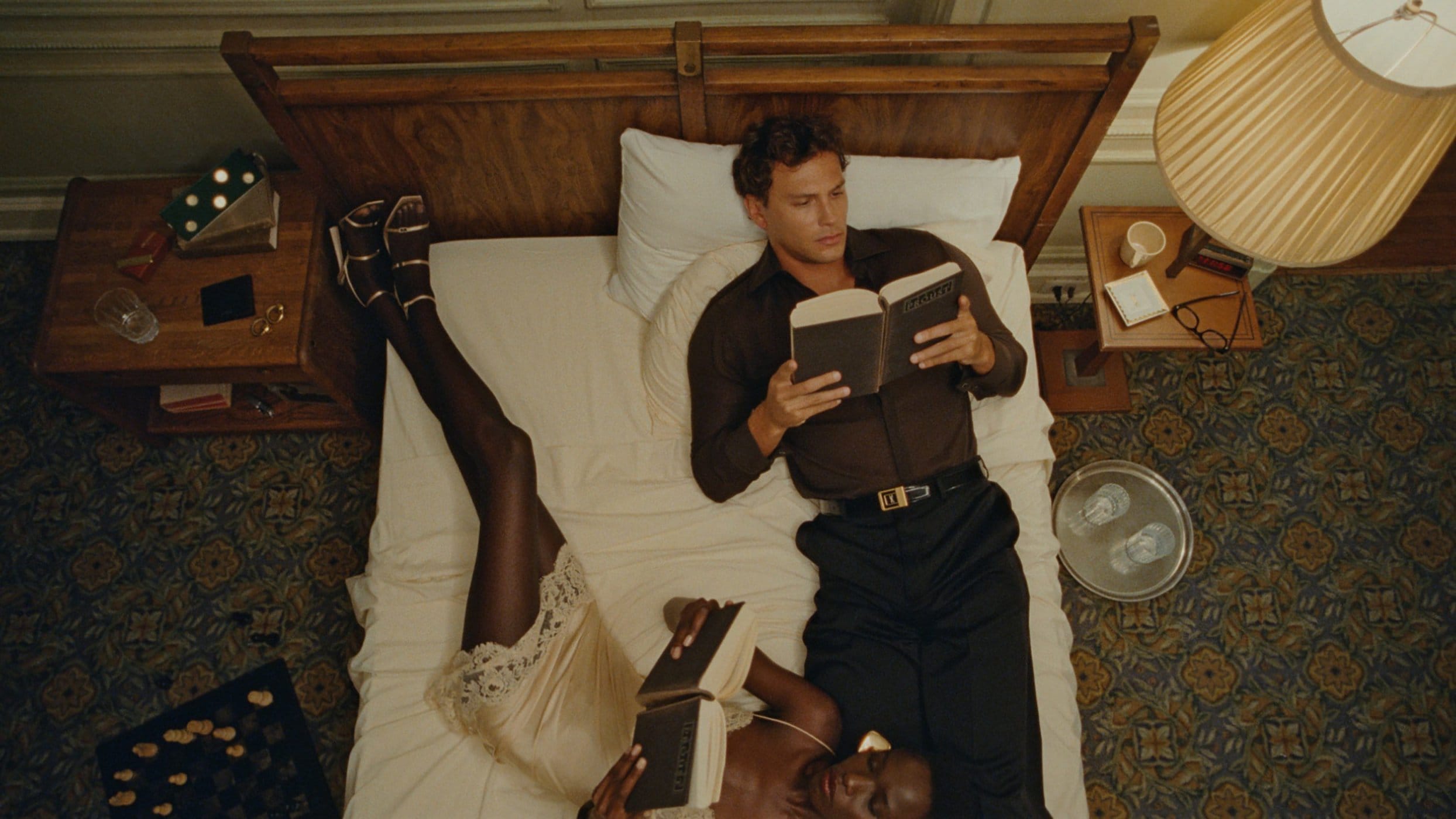 Man and woman lying on a bed reading books in a bedroom with patterned carpet and wooden nightstands