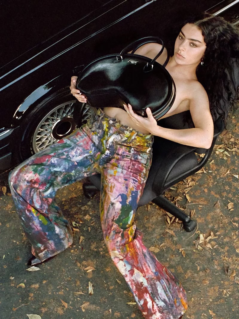 Charli XCX seated in a rolling office chair holding a black handbag in front of her chest wearing paint-splattered trousers
