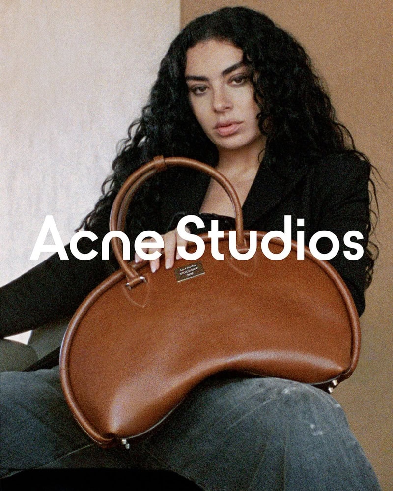 Woman with long dark curly hair wearing a black jacket holding a large brown leather handbag for Acne Studios Spring 2025 ad campaign.