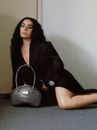 Woman in dark blazer and glasses posing on a grey floor next to a structured handbag