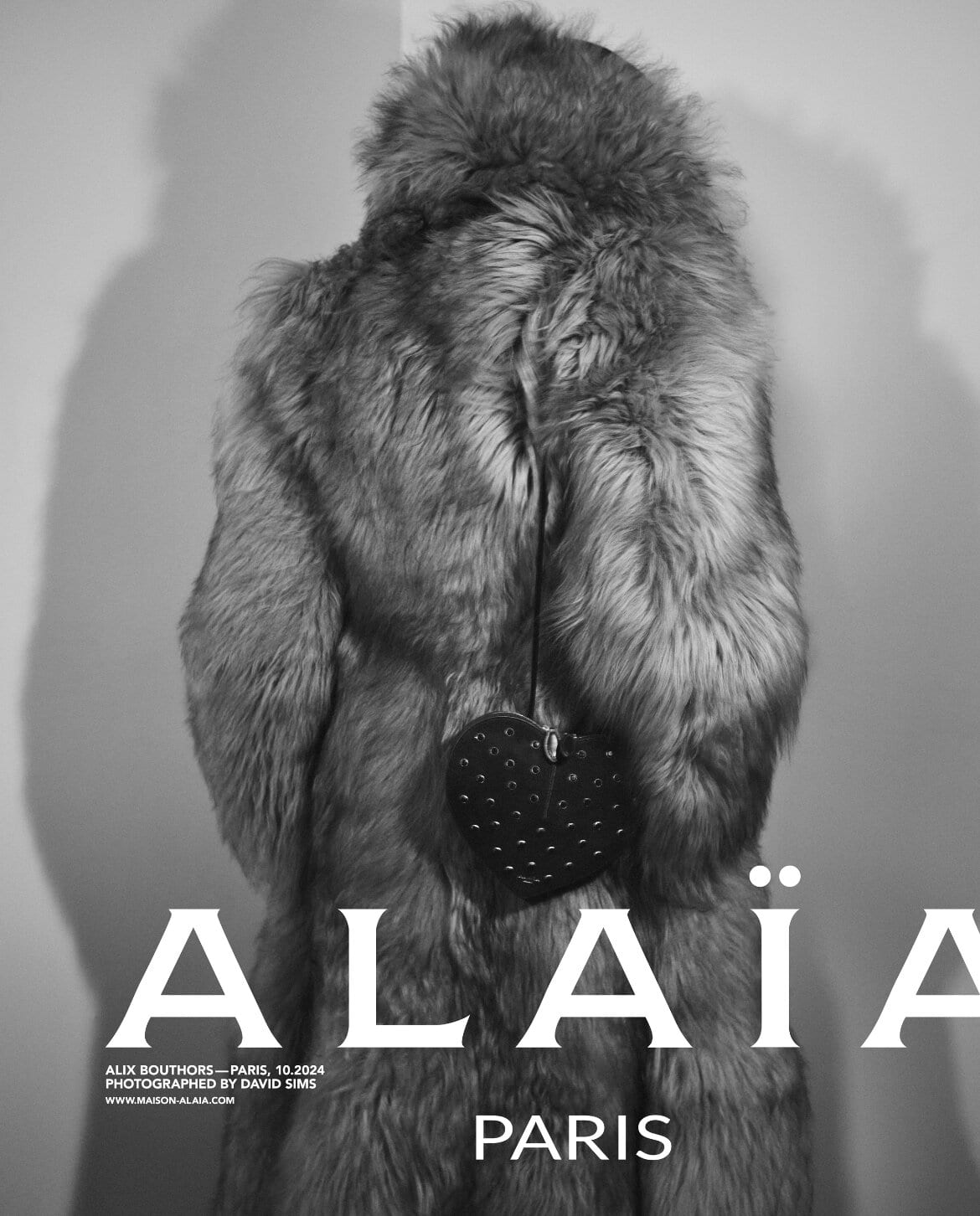 Model wearing a heavy textured fur coat from behind, featuring a heart-shaped bag against a plain wall