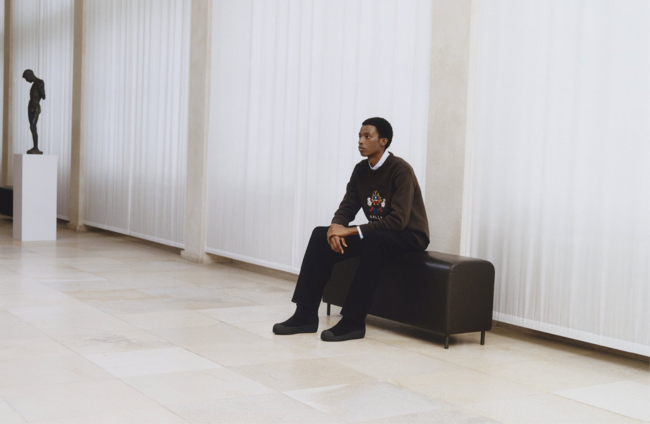 A young Black man seated on a dark bench inside a large hall with pale stone floors and tall white sheer curtains.