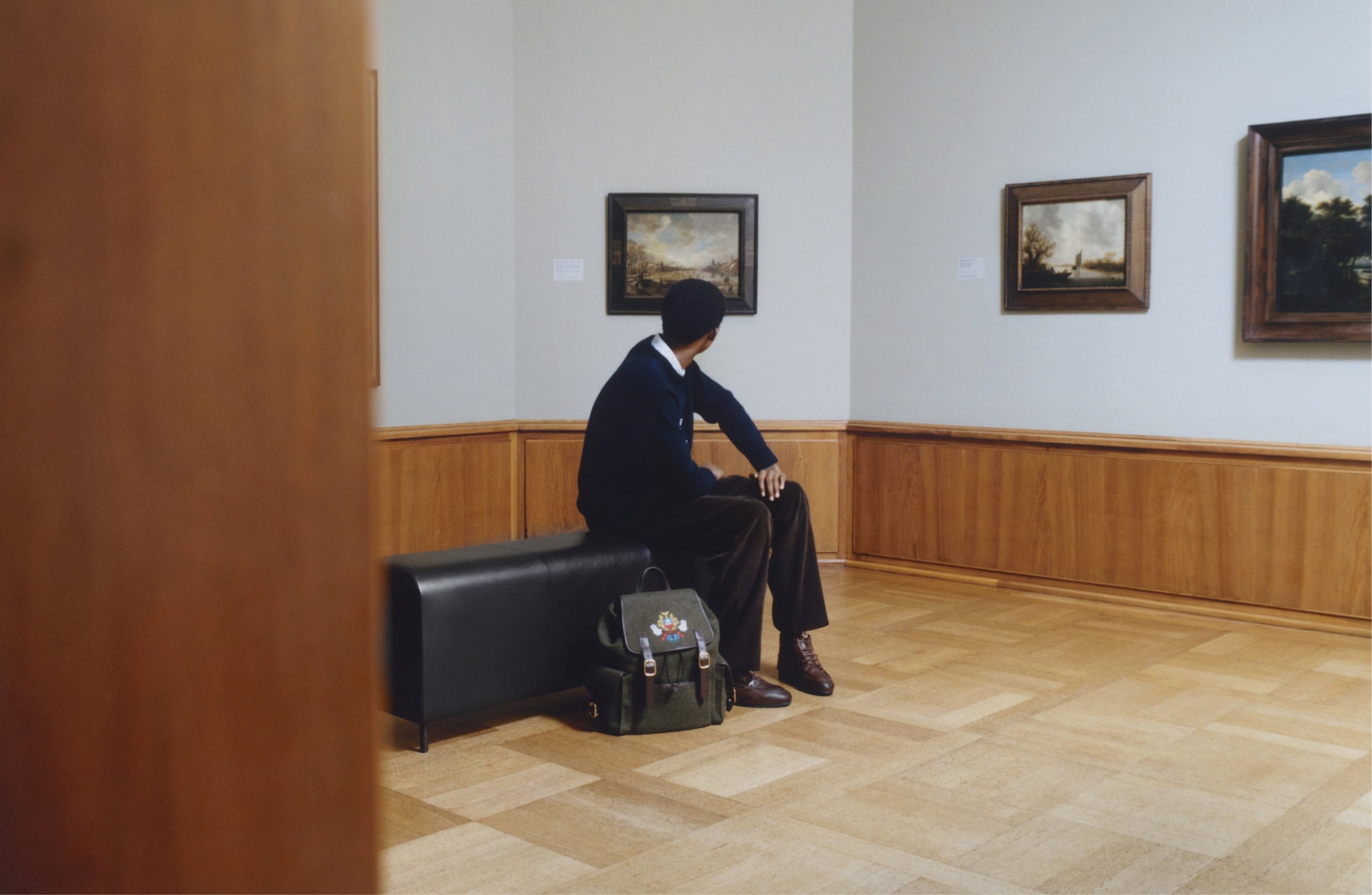 Man seated on a black bench in a museum gallery viewing framed landscape paintings on the wall.