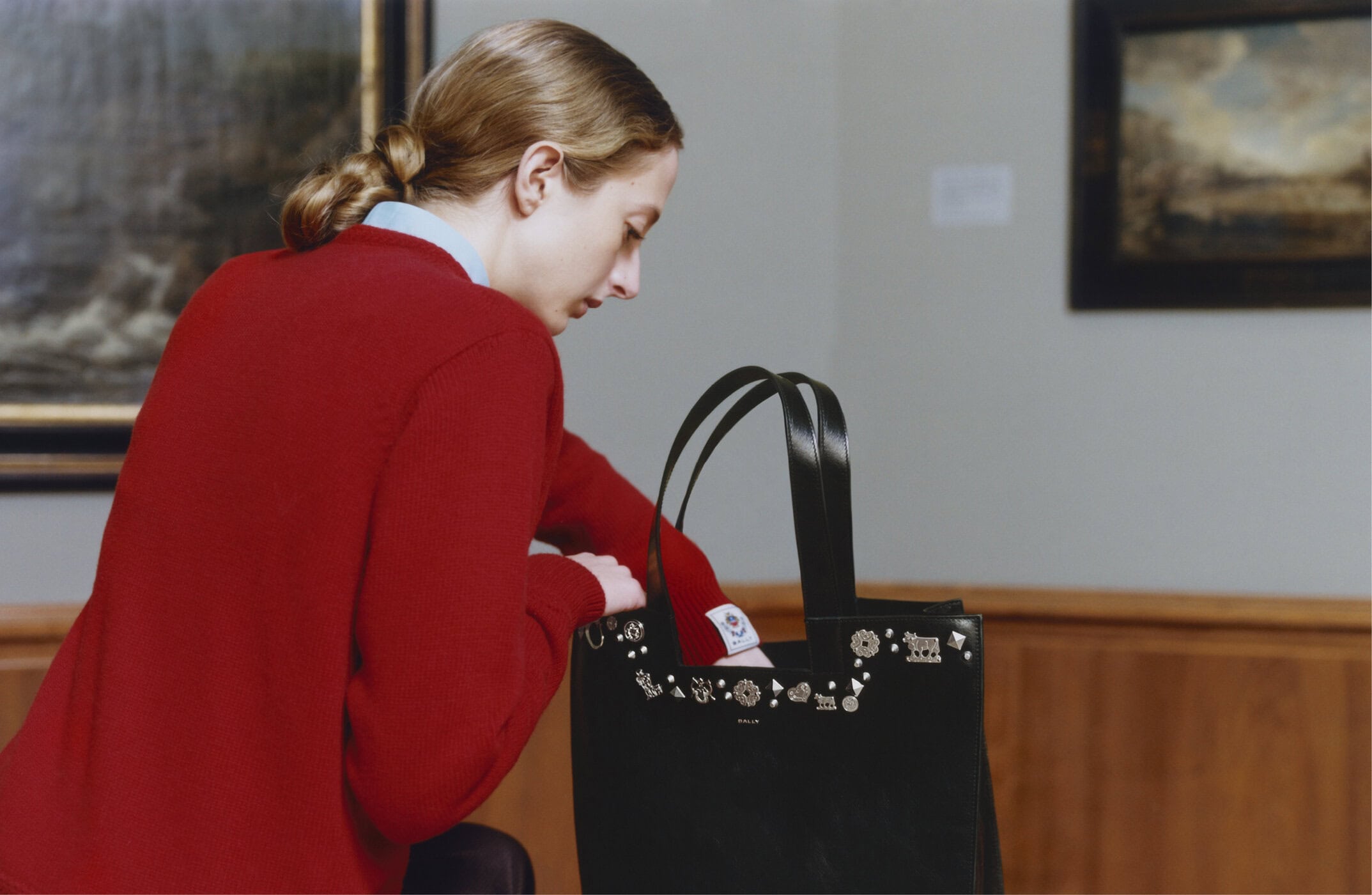 Woman in a red sweater reaching into a black leather tote bag adorned with metallic charms inside a museum gallery.