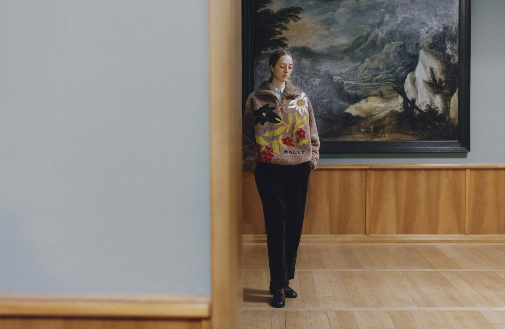 Model wearing a brown floral fleece pullover standing in an art gallery beneath a large landscape painting.