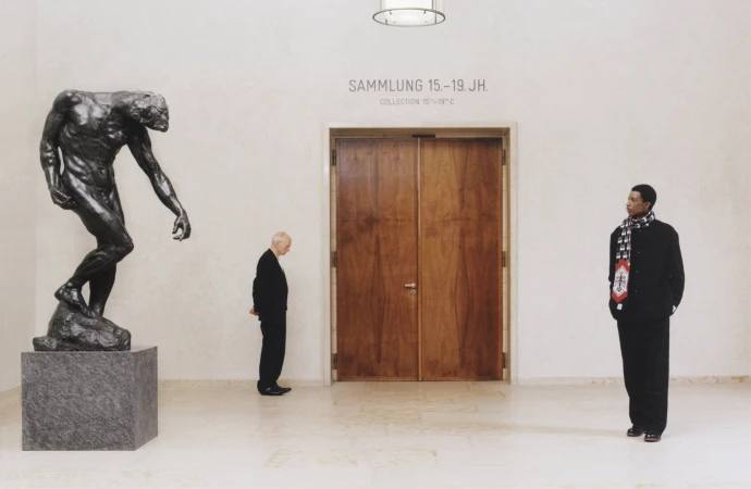 Man wearing black jacket and scarf standing inside museum gallery beside wooden double doors and bronze sculpture.