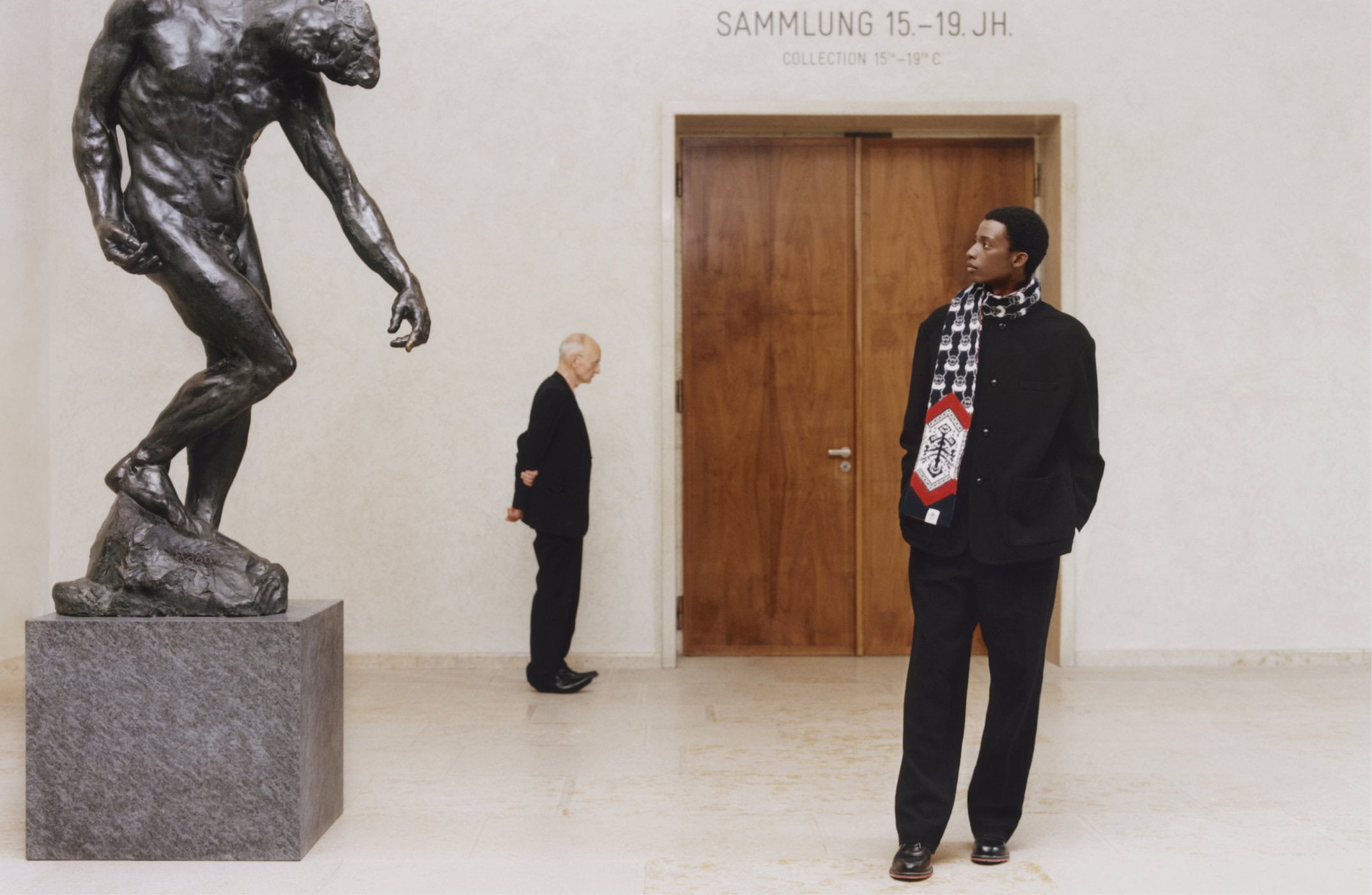 Man wearing a black jacket and patterned scarf standing in a gallery next to a large bronze sculpture.