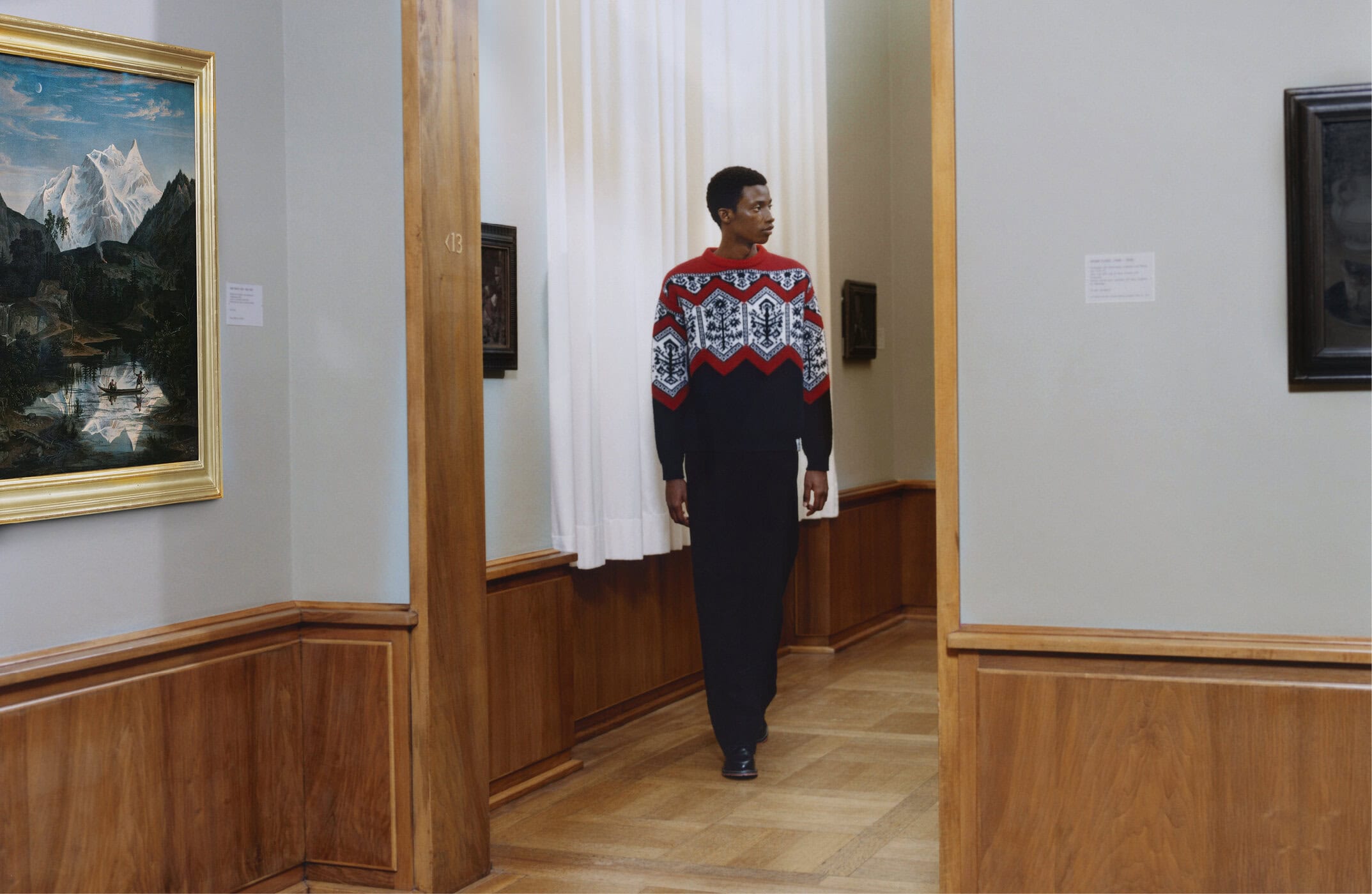 Man wearing a patterned red and white sweater and dark trousers standing inside an art gallery with framed paintings on the walls.