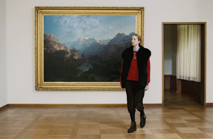 Model wearing a black fur vest, red sweater, and dark trousers standing inside a museum in front of a large landscape painting.