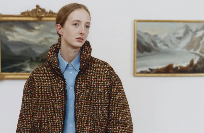 Model wearing patterned puffer jacket and blue button-down shirt inside a gallery with framed landscape paintings.
