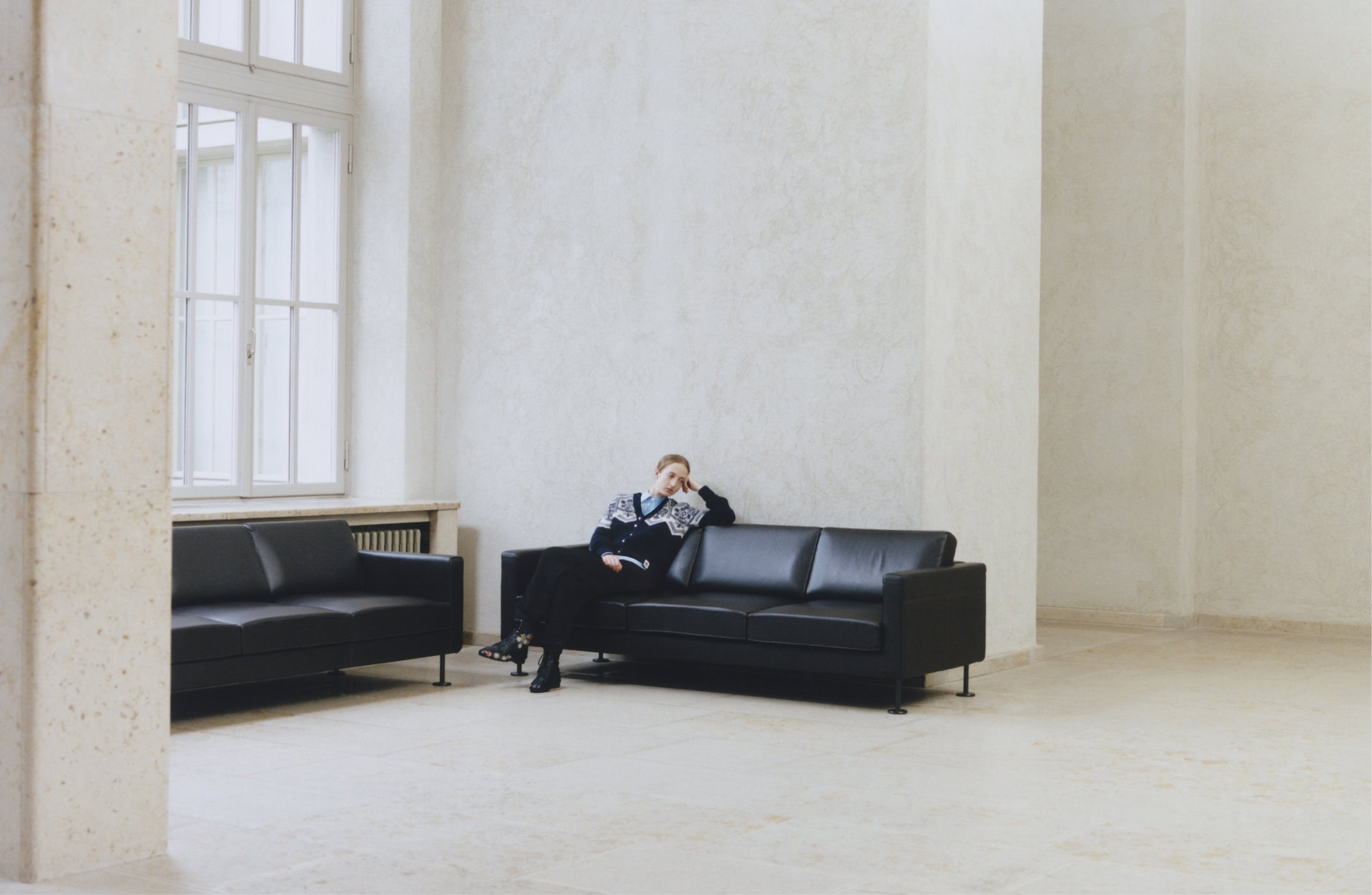 Model seated on a black leather sofa inside a grand light-filled hall with tall windows for Bally Winter 2024 campaign