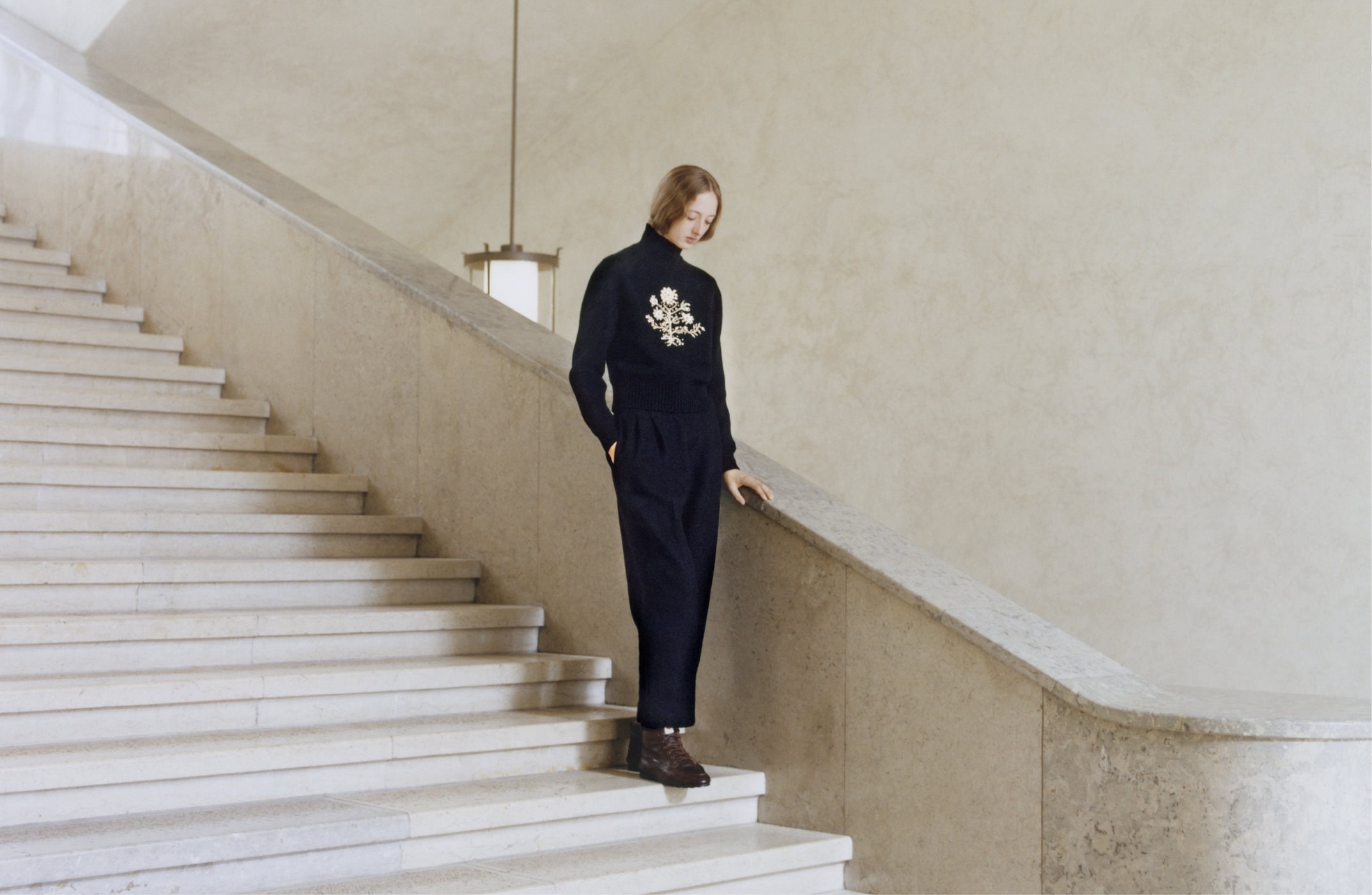 Model standing on a grand stone staircase wearing a dark turtleneck sweater with floral embroidery and dark trousers.
