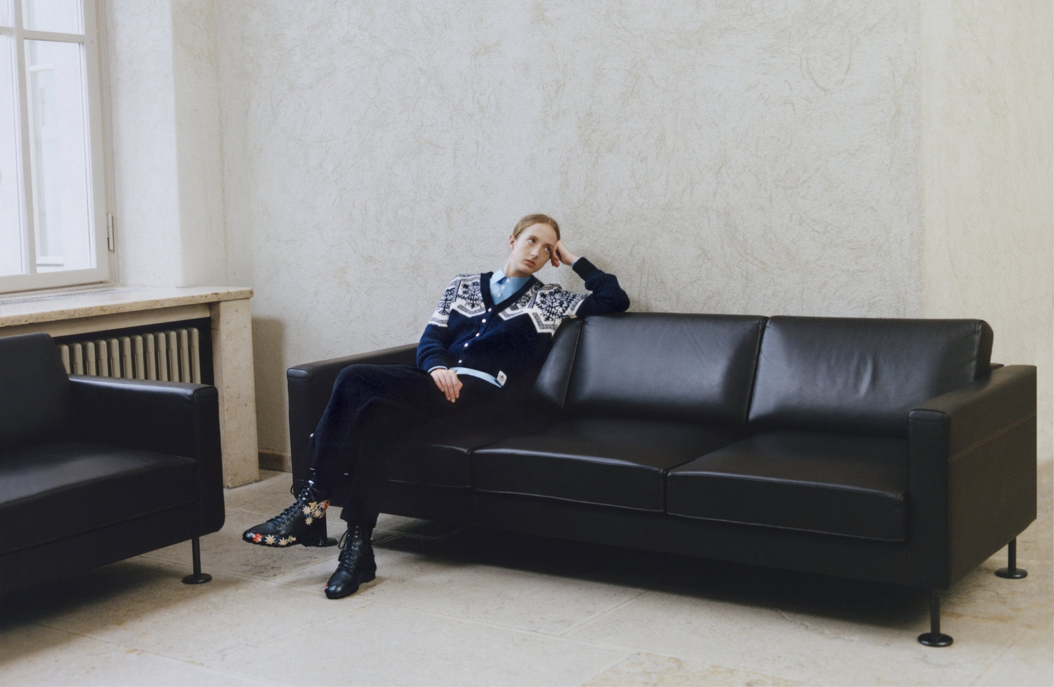Model wearing a patterned cardigan seated on a black leather sofa in an interior space with a window