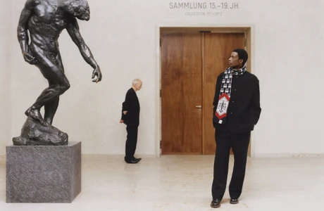 Man wearing a black jacket and patterned scarf standing in a gallery next to a large bronze sculpture.