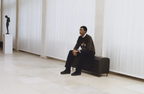 A young Black man seated on a dark bench inside a large hall with pale stone floors and tall white sheer curtains.