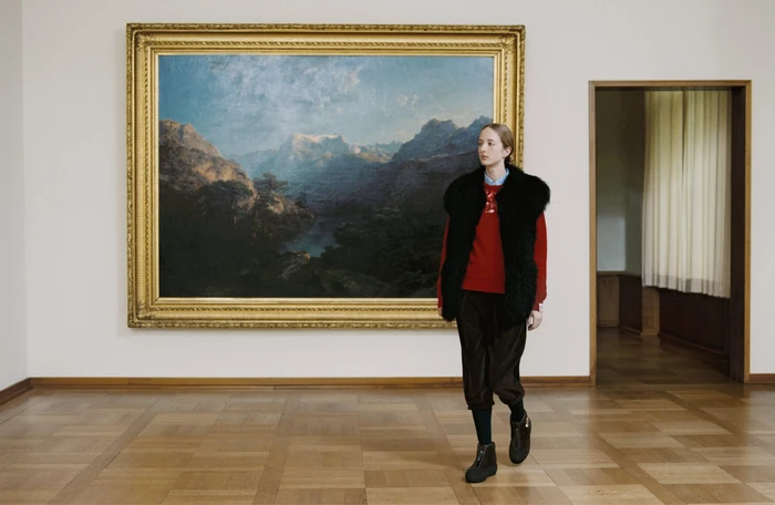 Model wearing a black fur vest, red sweater, and dark trousers standing inside a museum in front of a large landscape painting.