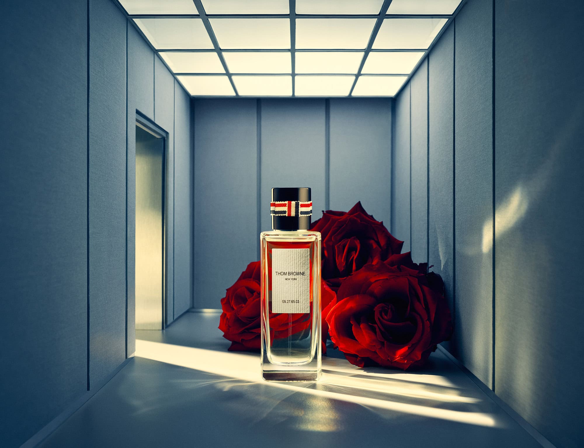 A rectangular glass perfume bottle with dark red roses behind it on a grey architectural floor under a gridded illuminated ceiling