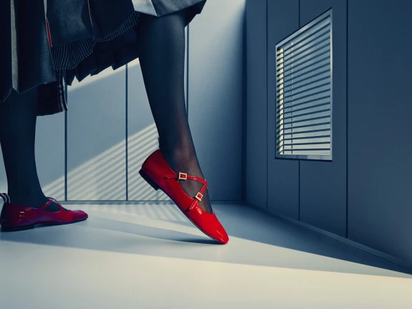 Close-up of legs in black hosiery and red patent leather Mary Jane flats stepping across a minimalist room with slatted window light.