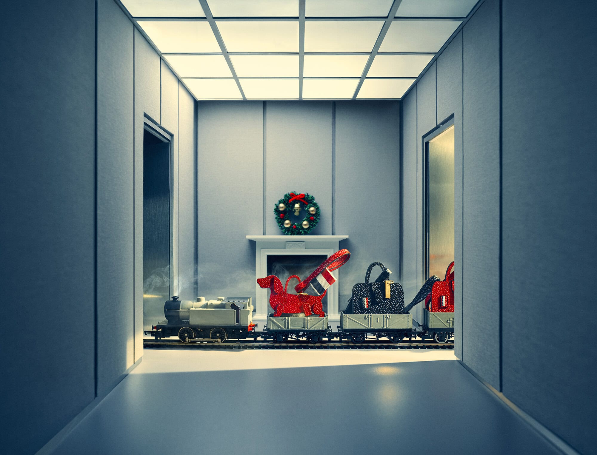 Toy train carrying luxury leather handbags and a dachshund bag on tracks through a grey interior hallway with a mantelpiece.
