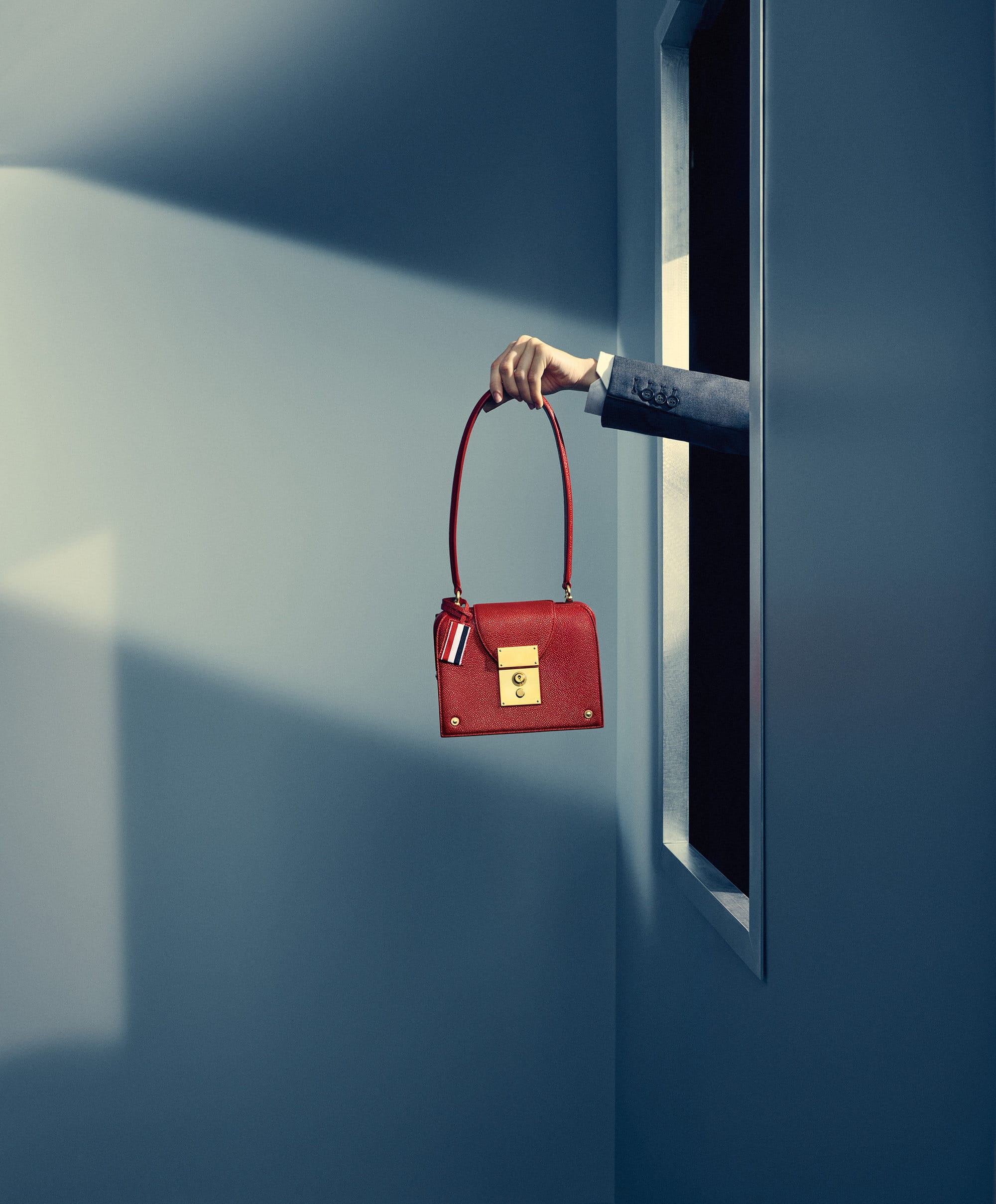 A hand extending from a vertical doorway holds a red leather handbag against a blue wall with dramatic lighting.