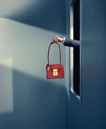 A hand extending from a vertical doorway holds a red leather handbag against a blue wall with dramatic lighting.
