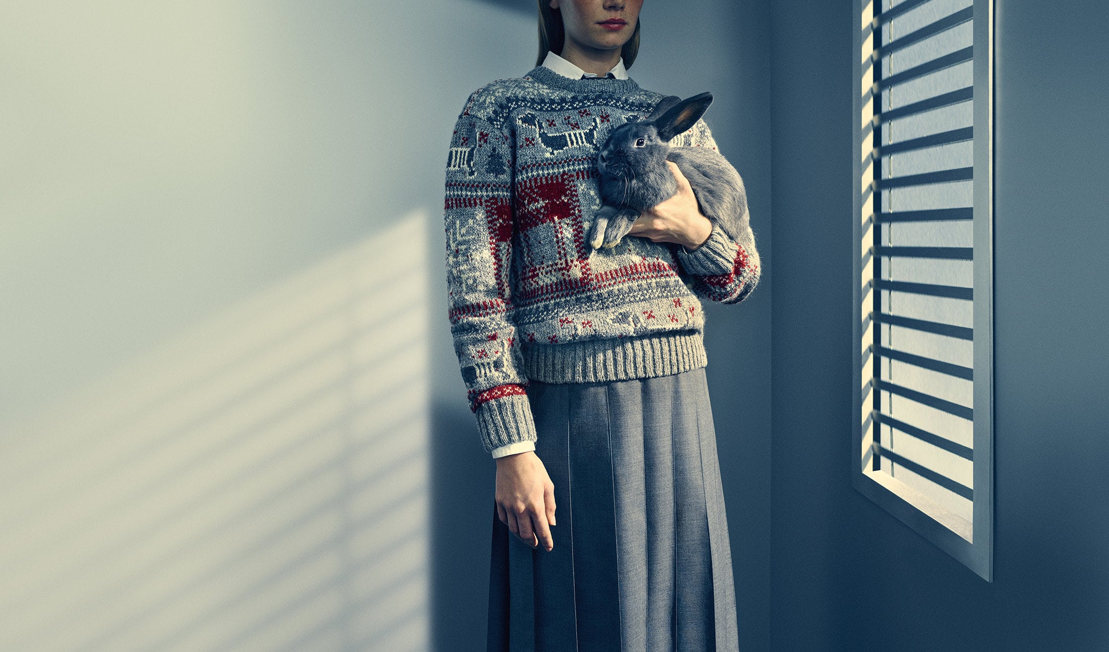 Model in grey patterned sweater holding a grey rabbit against a grey wall with window shadows