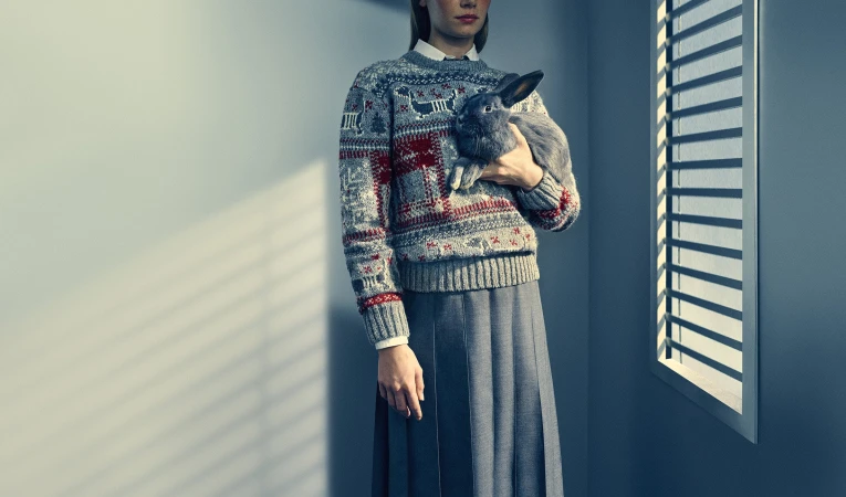 Model in grey patterned sweater holding a grey rabbit against a grey wall with window shadows