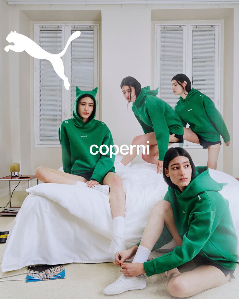 Model wearing green cat-eared hoodie and white sneakers posing on a white bed in a bright room for Coperni x Puma campaign