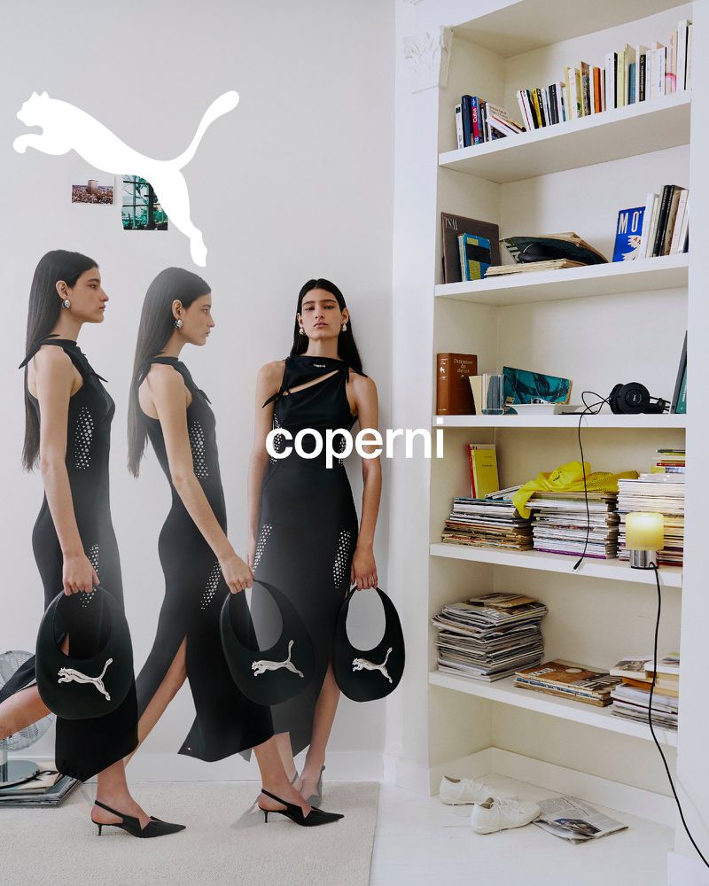 Model in a black dress holding a curved handbag against a white wall beside a white bookcase