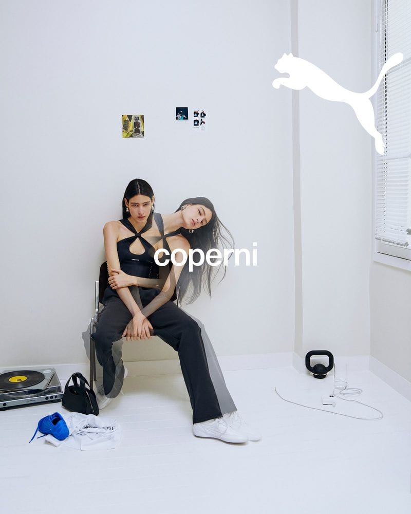 Model seated on a chair in a white room wearing a black top and trousers with white sneakers for Coperni x Puma Fall 2024