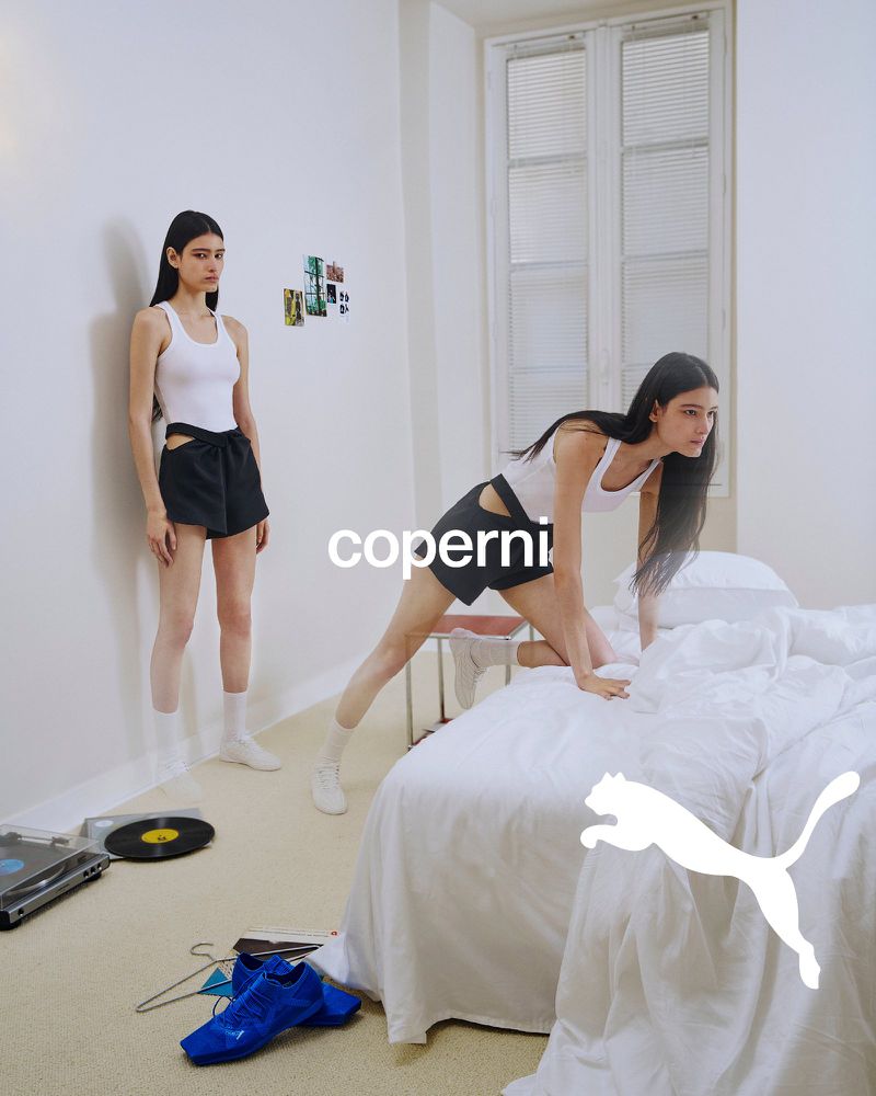 Two female models in white tank tops and black shorts inside a sunlit bedroom setting with blue sneakers on the floor.