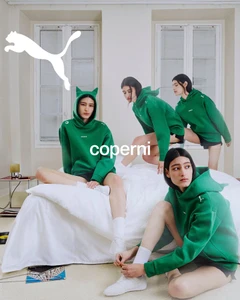 Model wearing green cat-eared hoodie and white sneakers posing on a white bed in a bright room for Coperni x Puma campaign