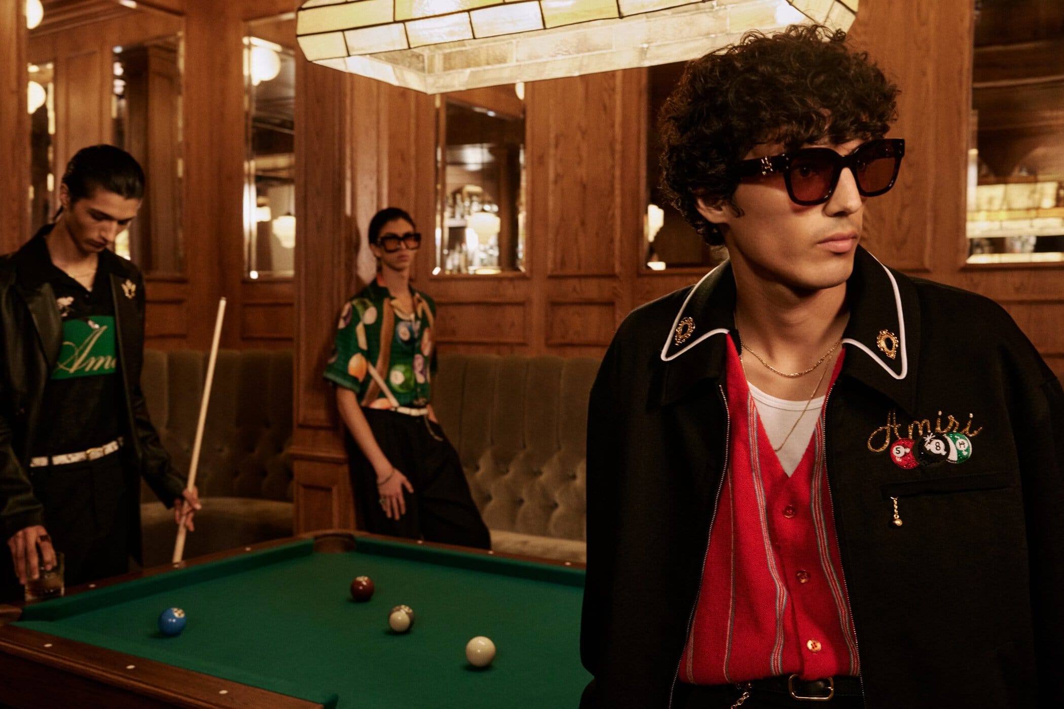 Three male models in a wood-paneled billiard room with a green pool table for the Amiri Pre-Spring 2025 ad campaign.