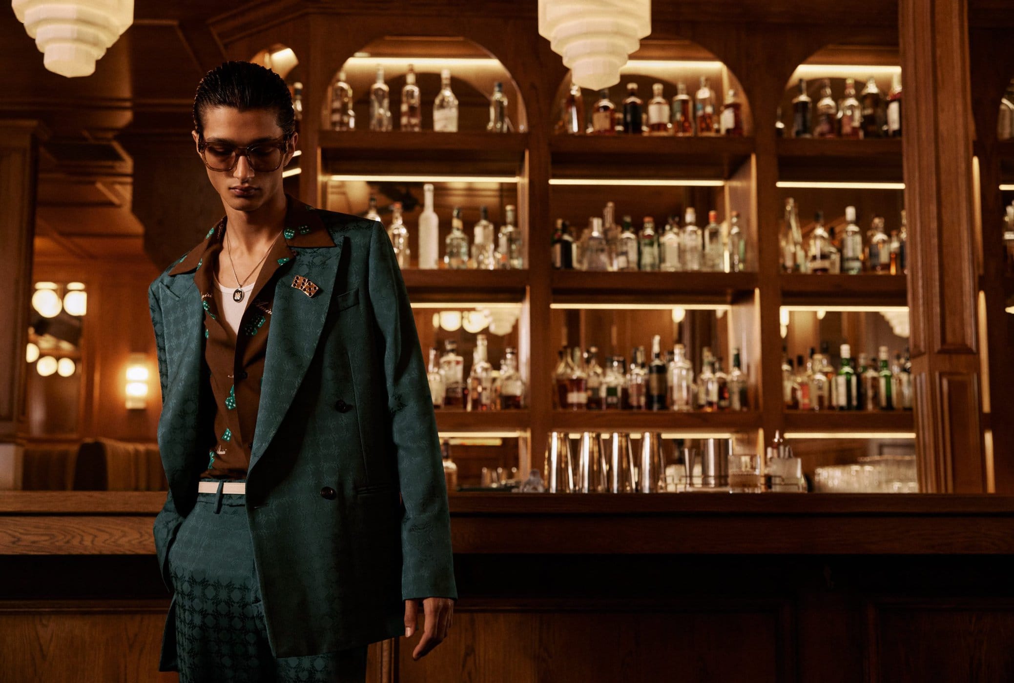 Man in a dark teal patterned suit and brown shirt standing inside a bar with illuminated shelves of bottles