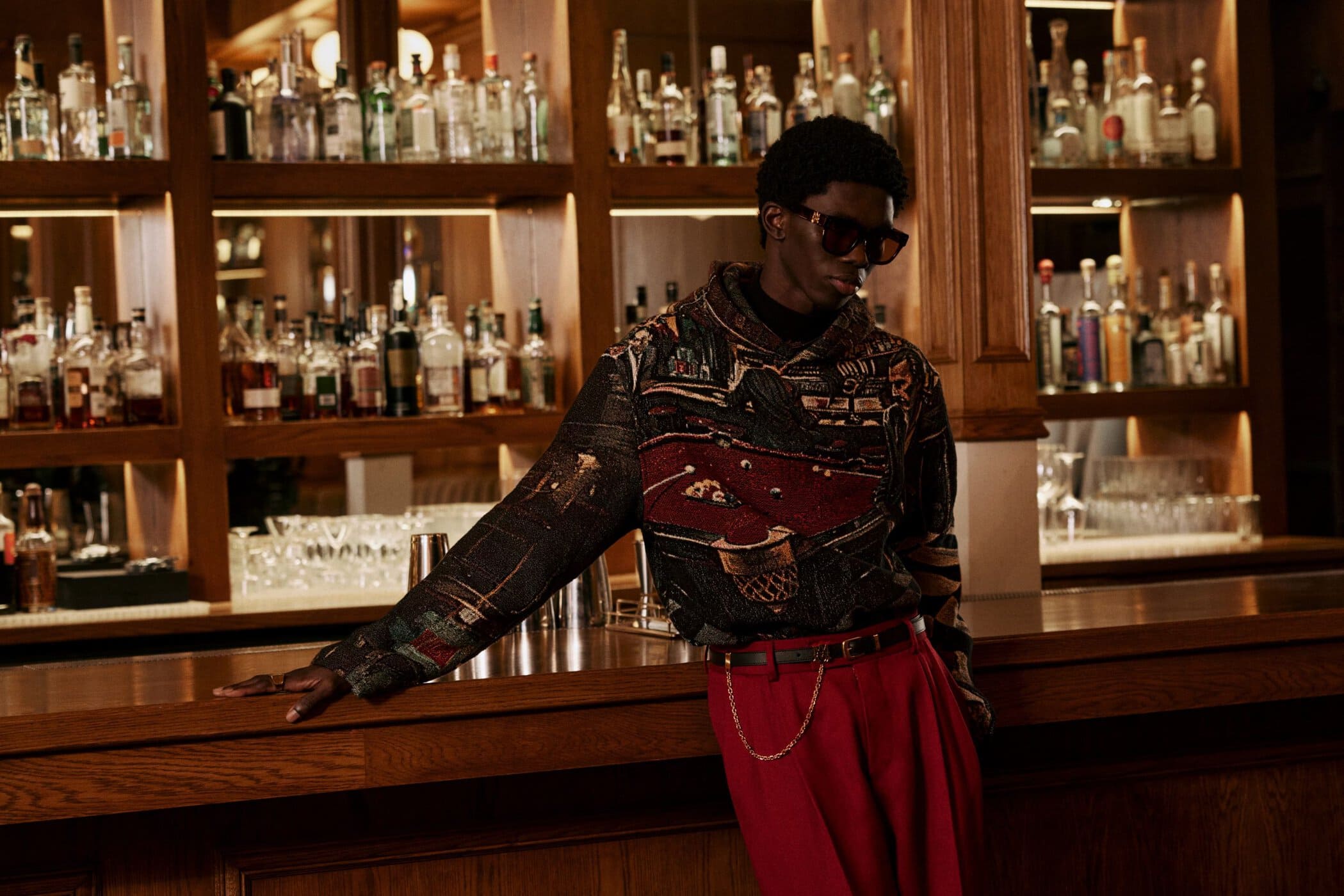 Model wearing patterned hoodie and red trousers leaning against a wooden bar in a dimly lit lounge