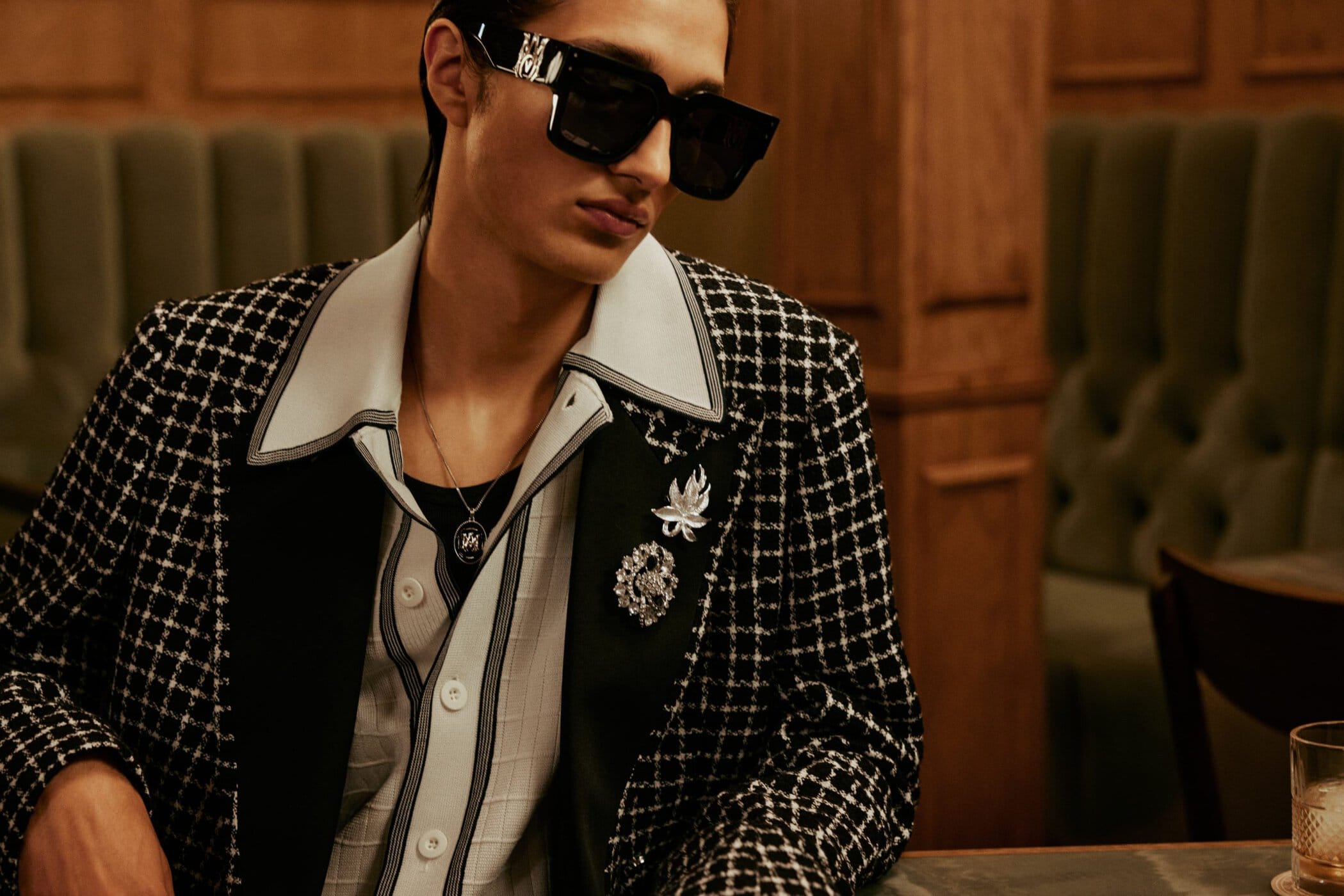 Man wearing a black and white houndstooth jacket, white collared shirt, and black sunglasses in an interior setting.