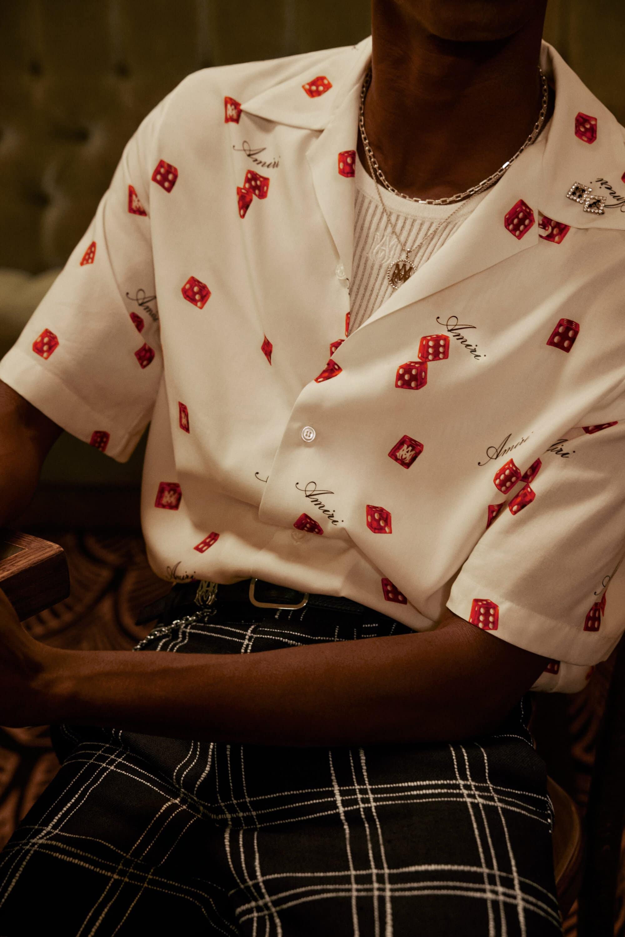 Close-up of a person wearing a white short-sleeve button-up shirt with a red dice pattern and dark plaid trousers, accessorized with silver necklaces.