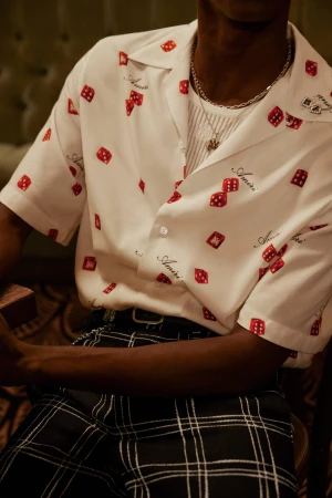 Close-up of a person wearing a white short-sleeve button-up shirt with a red dice pattern and dark plaid trousers, accessorized with silver necklaces.